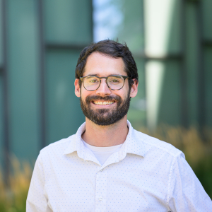 Smiling headshot of award winner Adam Felts in front of a green and black background wearing an off white button down long sleeve shirt.