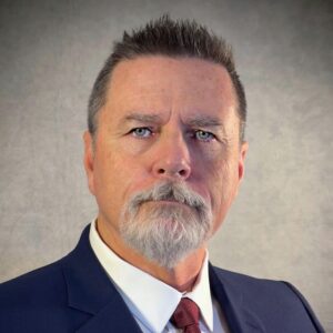 This is the headshot of an award winner looking serious with a grey goatee and dark colored suit and red tie in front of a dark grey background