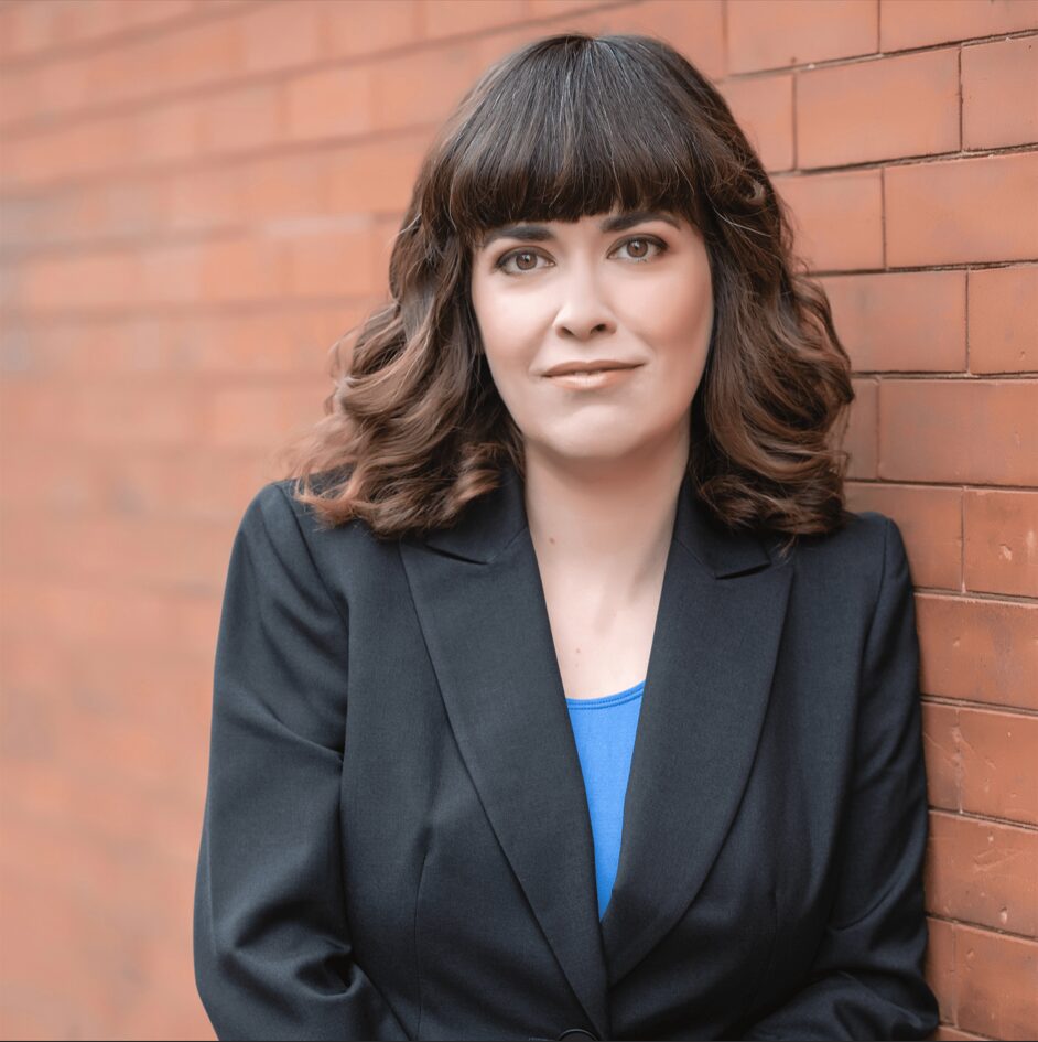 maroon brick background, white woman standing in front of it, dark blazer, blue shirt, long brown hair