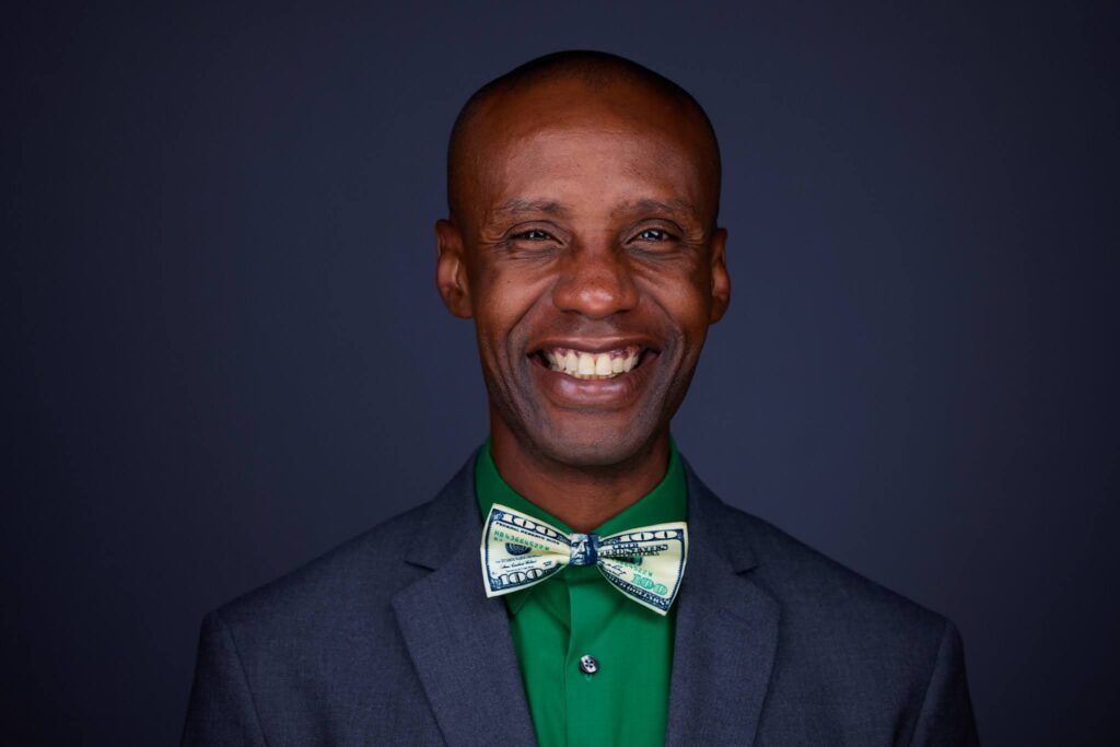 Dark background, man of color, light colored bow tie, green shirt, dark blazer