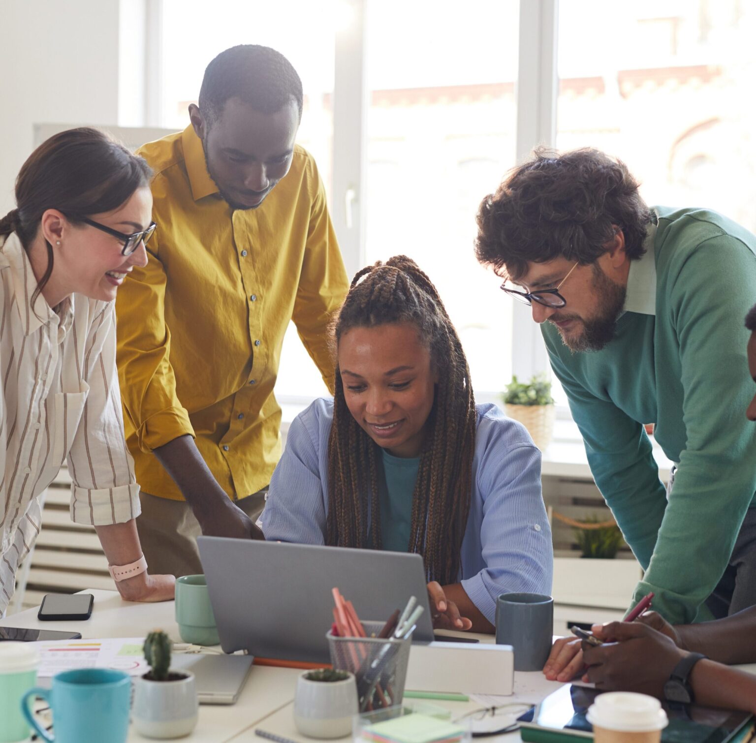 A diverse group of five people collaborating around a laptop in a bright, modern office space. The group appears engaged, with one person typing while others observe and contribute. The table is cluttered with notebooks, pens, and coffee cups, emphasizing a casual yet productive work environment.